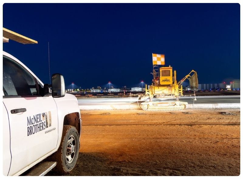 A white truck with the McNeil Brothers logo on the side parked near a paving machine