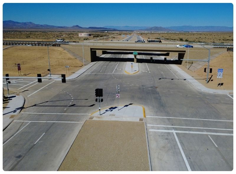 High-angle, eye-level aerial view of a wide, newly paved concrete roadway in Arizona leading to an overpass under a clear, bright blue sky in a desert landscape.