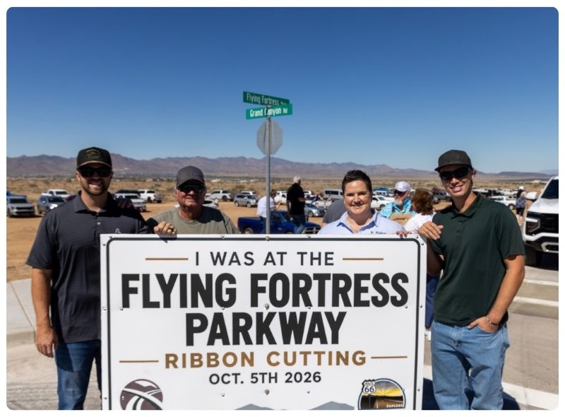 Four individuals pose outdoors under a clear blue sky, holding a large white sign that reads: