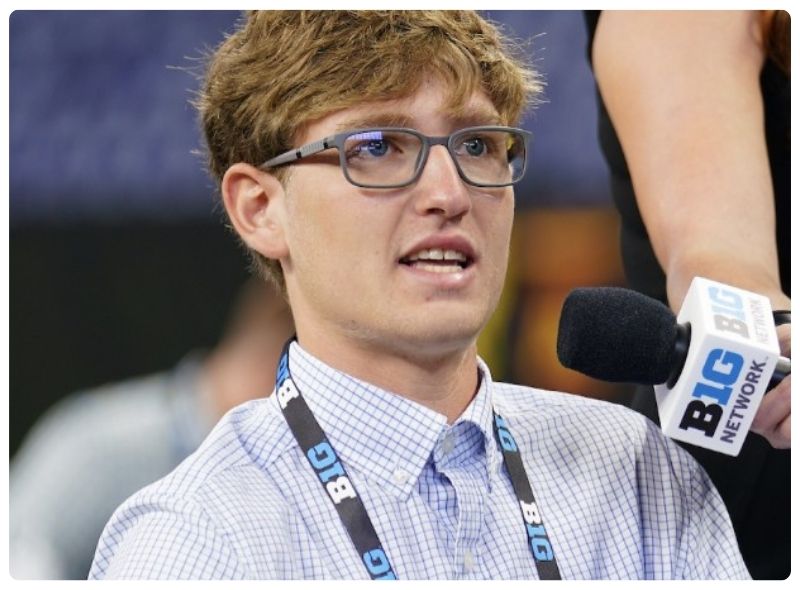 A person with light brown hair and glasses is speaking into a microphone labeled 