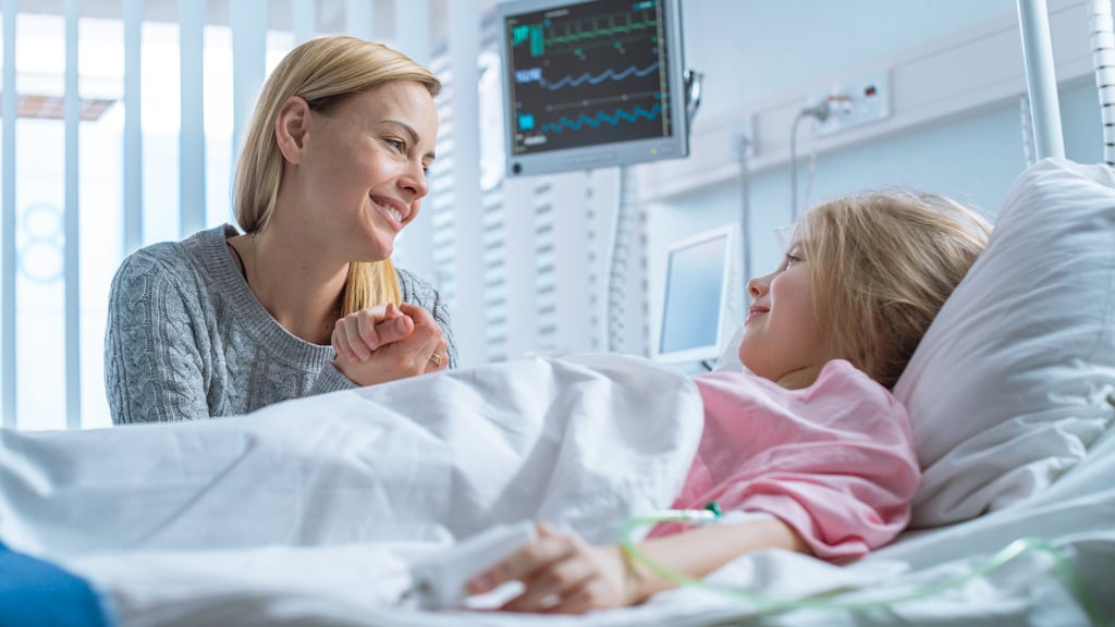 Visiting mother holding hand of young smiling daughter laying in a hospital bed