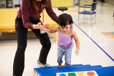 A physical therapist helps a toddler-aged girl climb stairs