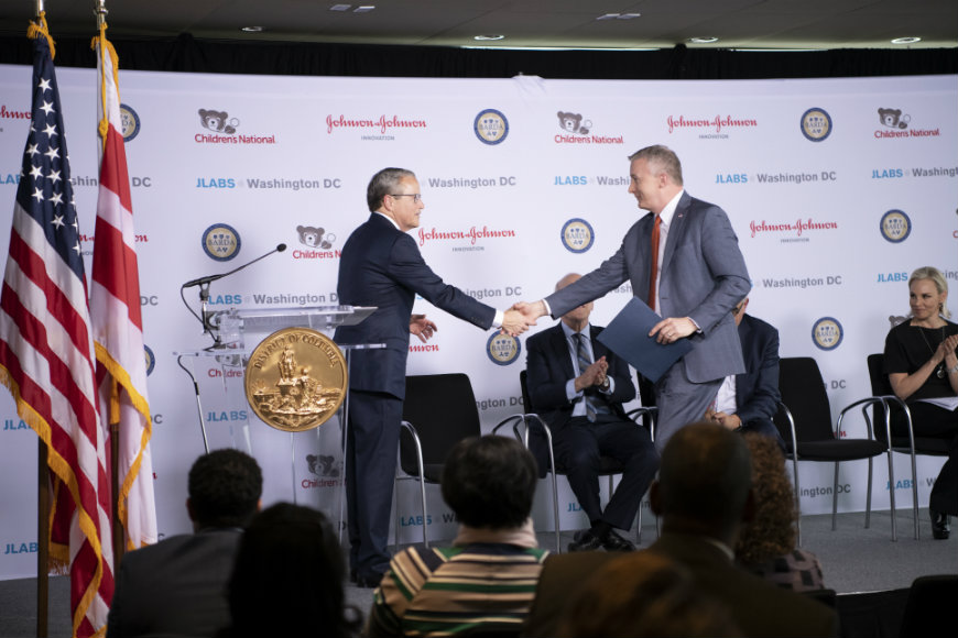 Two men shake hands on the stage during the press conference; the American and District of Columbia government flags are in the background.