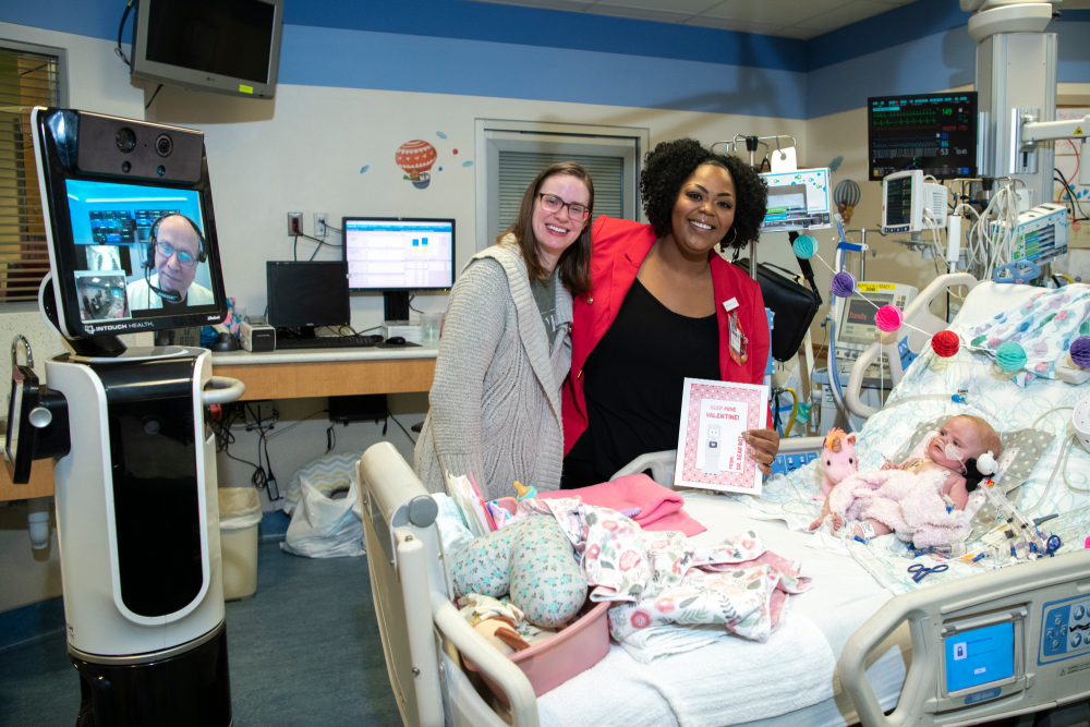 dr. bear bot, dr. lopez and monique hold valentine card for infant on a hospital bed