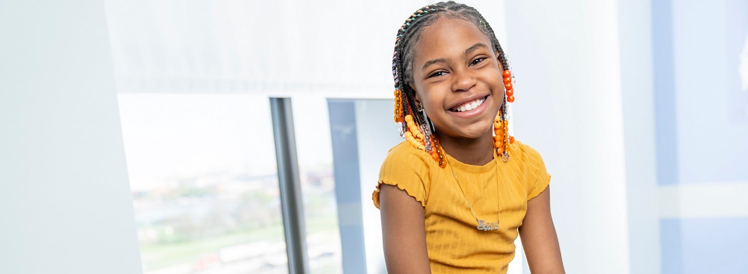 A teen girl in an orange shirt smiles as she waits to see her doctor at a primary care visit.