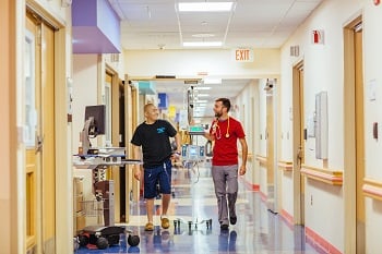 A male nurse helps an adolescent male patient walk down a hospital hallways while the patient is hooked up to a tall wheeled monitor. 