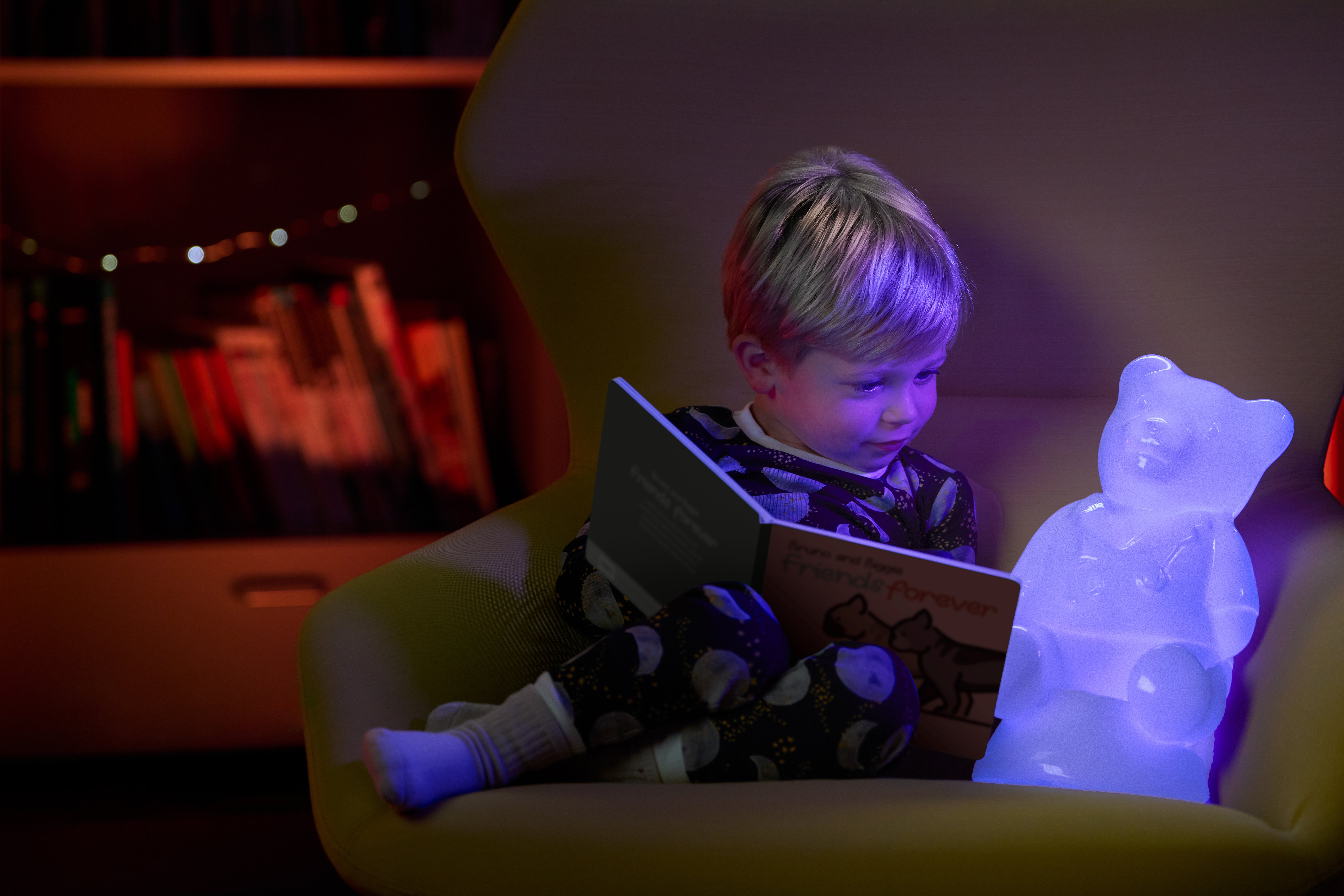 A White male toddler reads a bedtime story in a big chair with a lit up Dr. Bear alongside him.