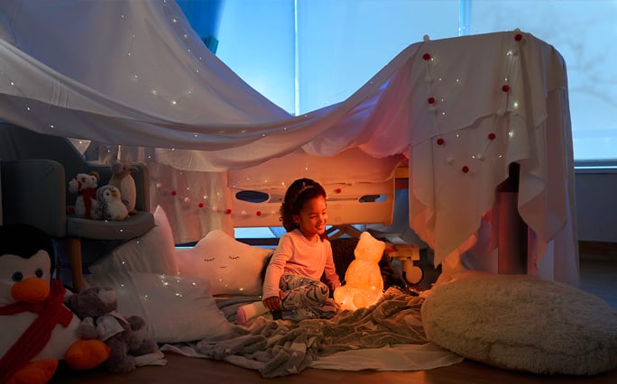 An elementary school-aged Black female patient sits in the warm glow of her hospital room "fort" lit up by Dr. Bear.