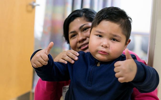 A male Latino toddler sits on his mother's lap and gives two thumbs up.