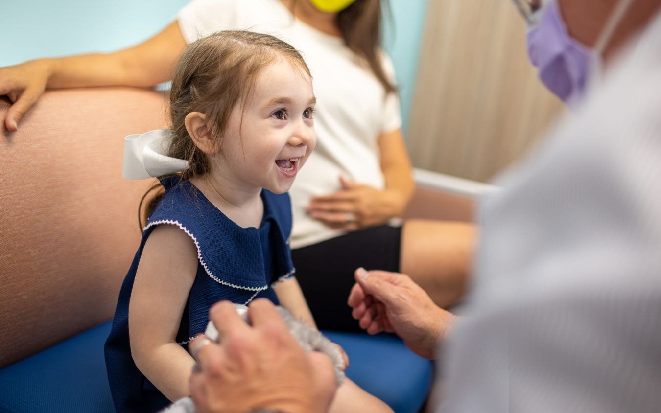 A White pre-school aged girl sits on a couch next to her mother while a practitioner engages her.