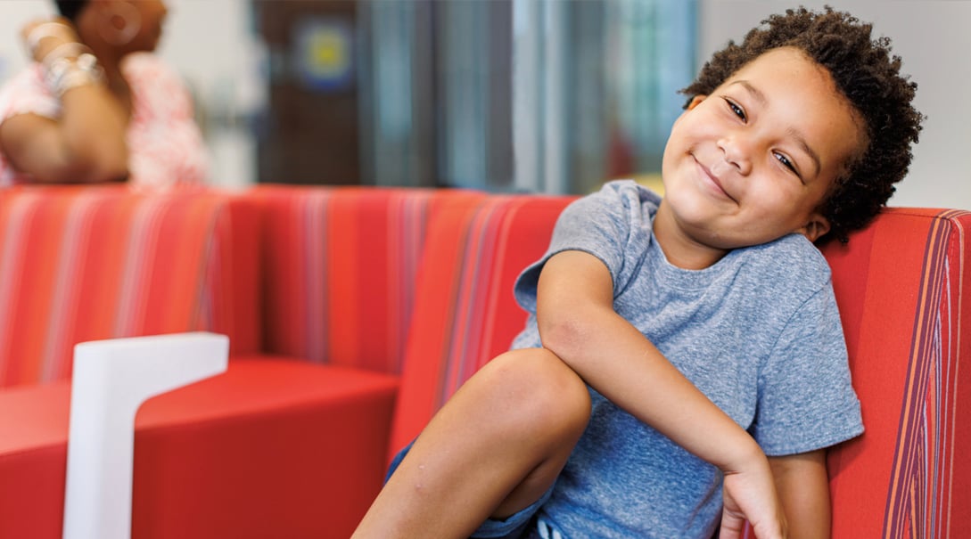 A Black pre-school aged male patient smiles in a hospital lounge.