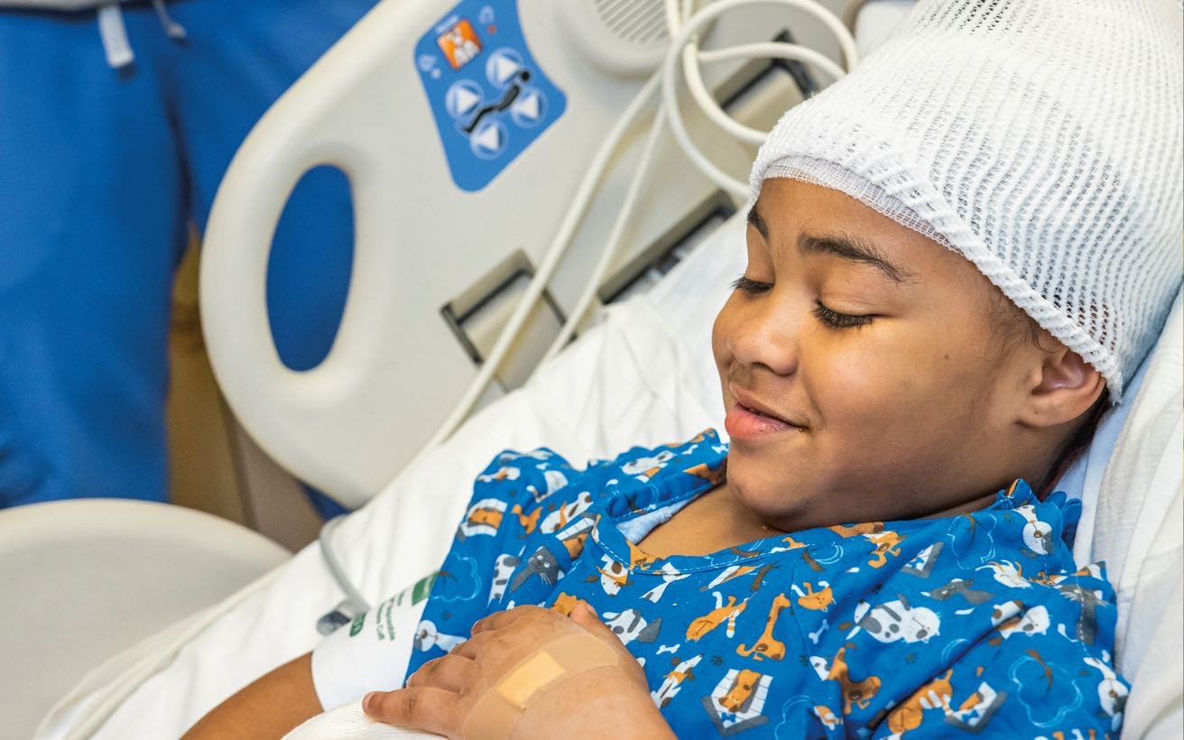 An Black elementary school-aged patient wearing a head bandage recovers in their hospital bed.