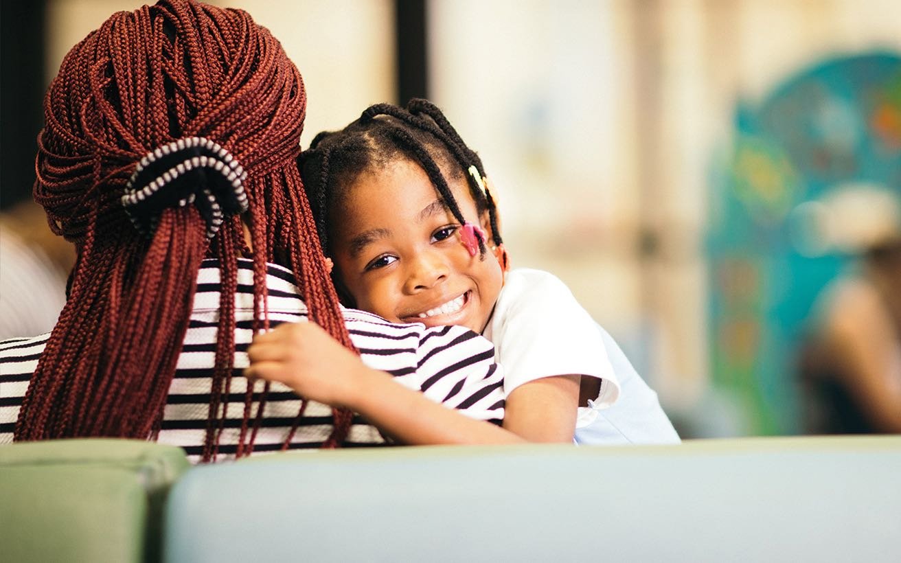 A Black mother sits in a waiting room holding her pre-school aged child.