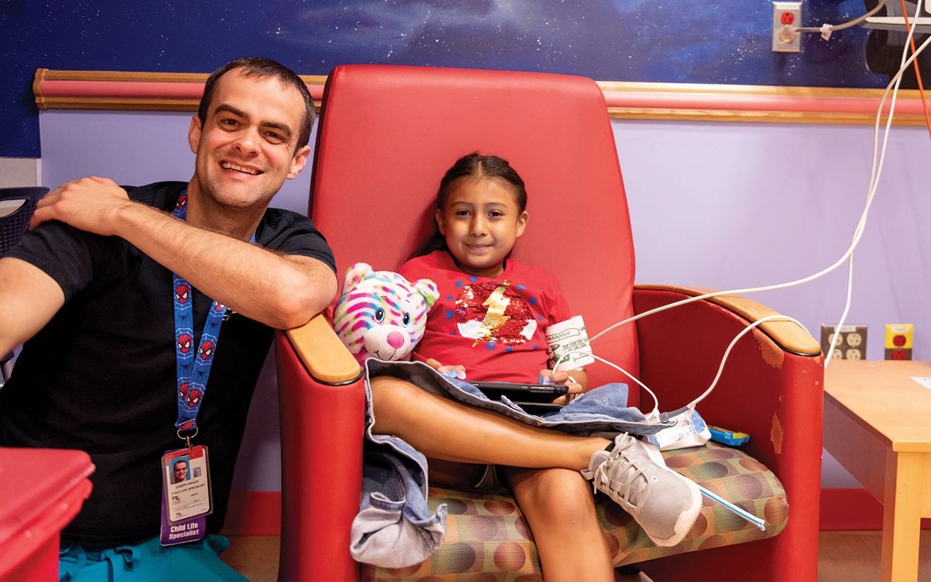 A male Child Life Specialist kneels next to a female school aged-patient undergoing chemotherapy treatment.