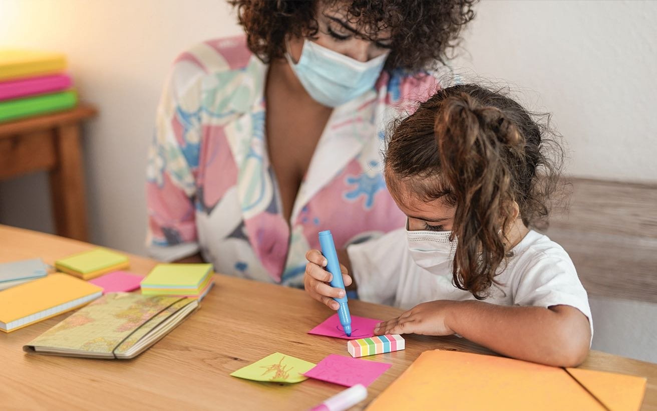 A pre-school aged female child uses markers to color. An adult female sits with her at the table.