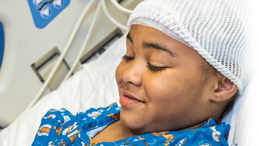 An elementary-school aged patient of color smiles in their hospital bed post surgery.