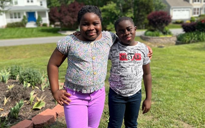 Two Black children, a girl and a boy, pose for a portrait with arms around each other on the front lawn of their home.