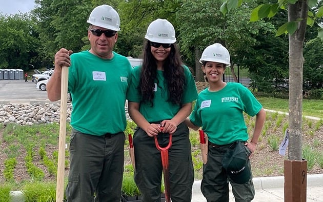 Three people posing in hardhats and t-shirts with landscape tools.