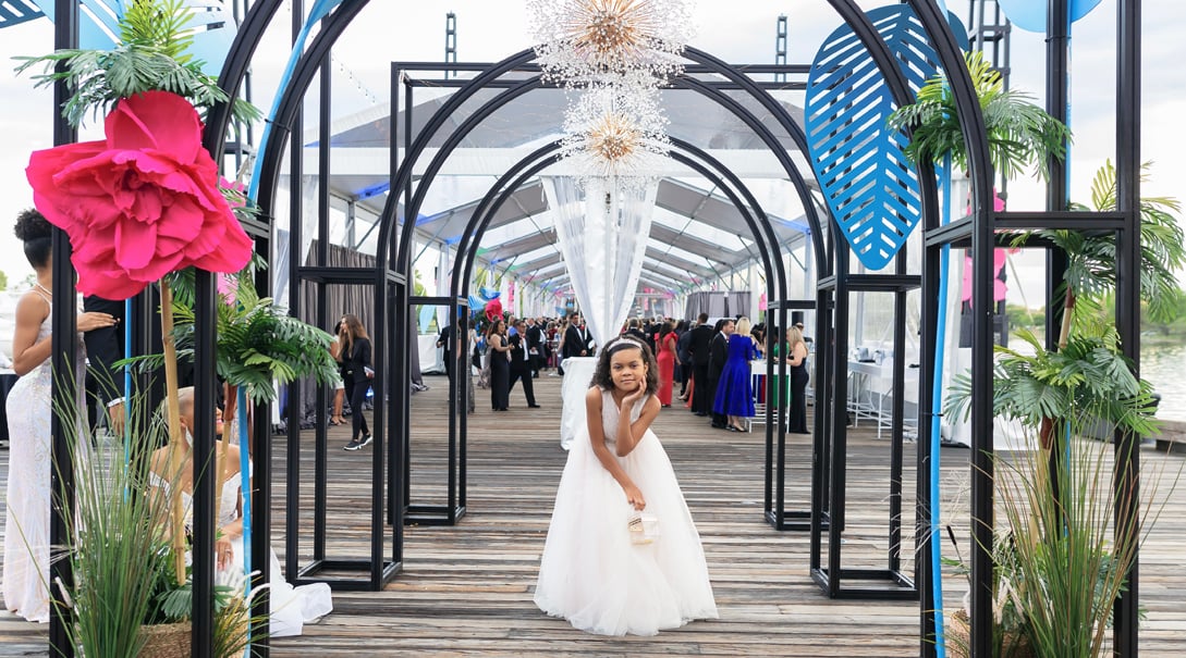 A Black pre-teenaged female wearing a formal white gown poses for a portrait at a gala event.