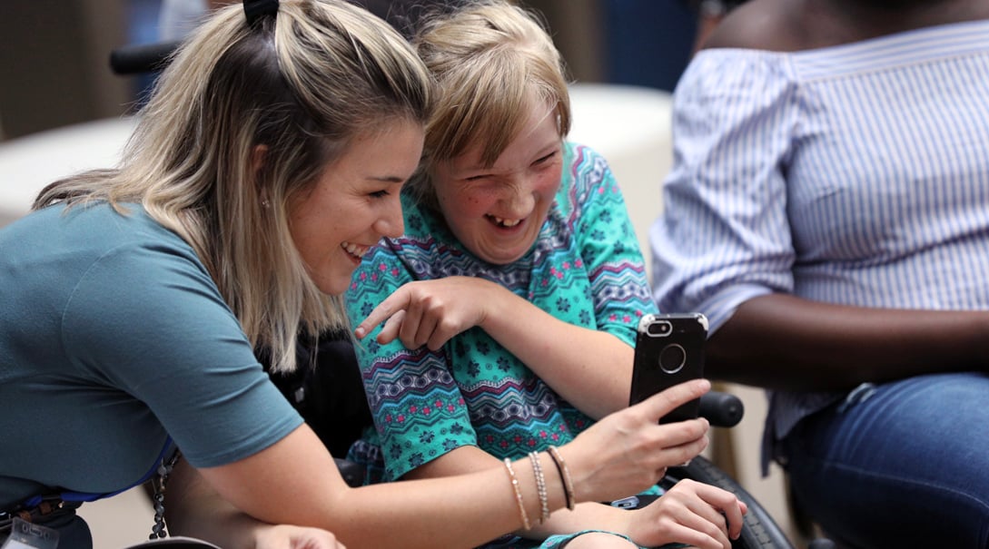 A White female patient in a wheelchair shares a laugh with a White woman while looking at the woman's smartphone.