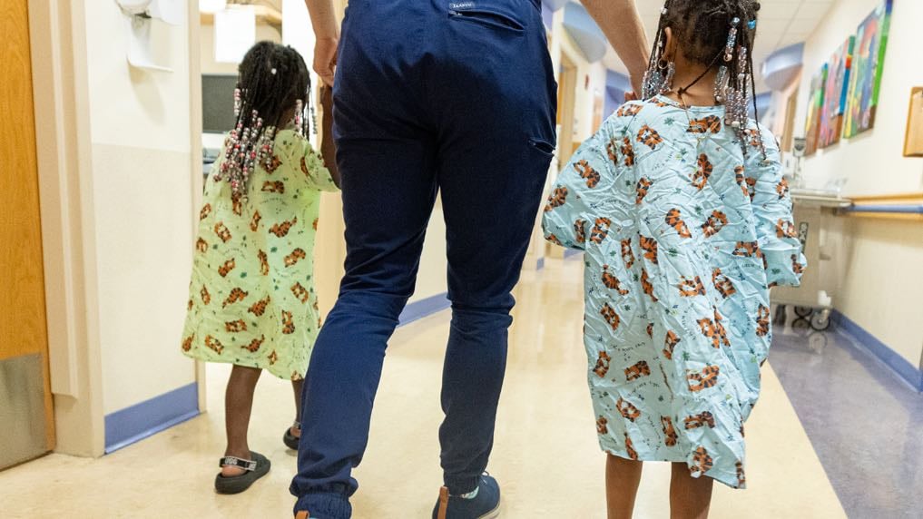 A rearview shot of a hospital practitioner walking hand-in-hand with two elementary school aged Black girls.