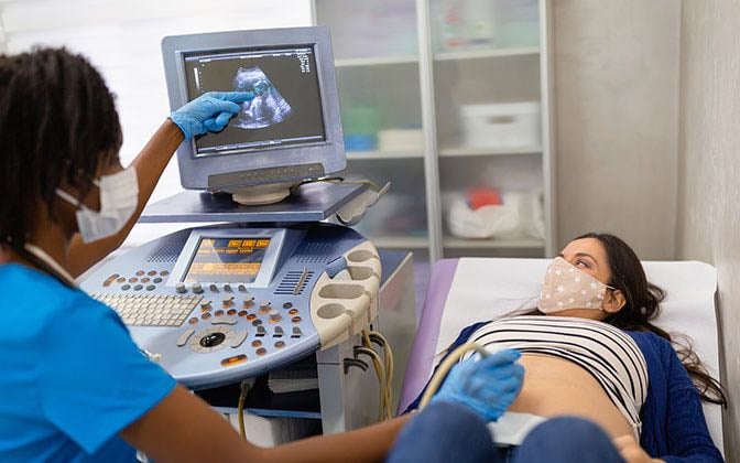 A Black female imaging technician explains what she is seeing on the monitor to an expectant mother who is White.