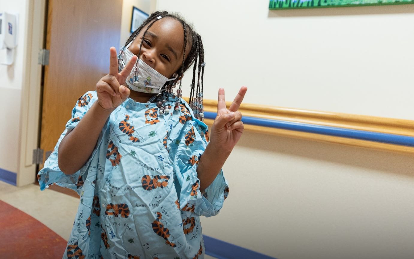 A Black elementary school aged female patient gives the peace sign.