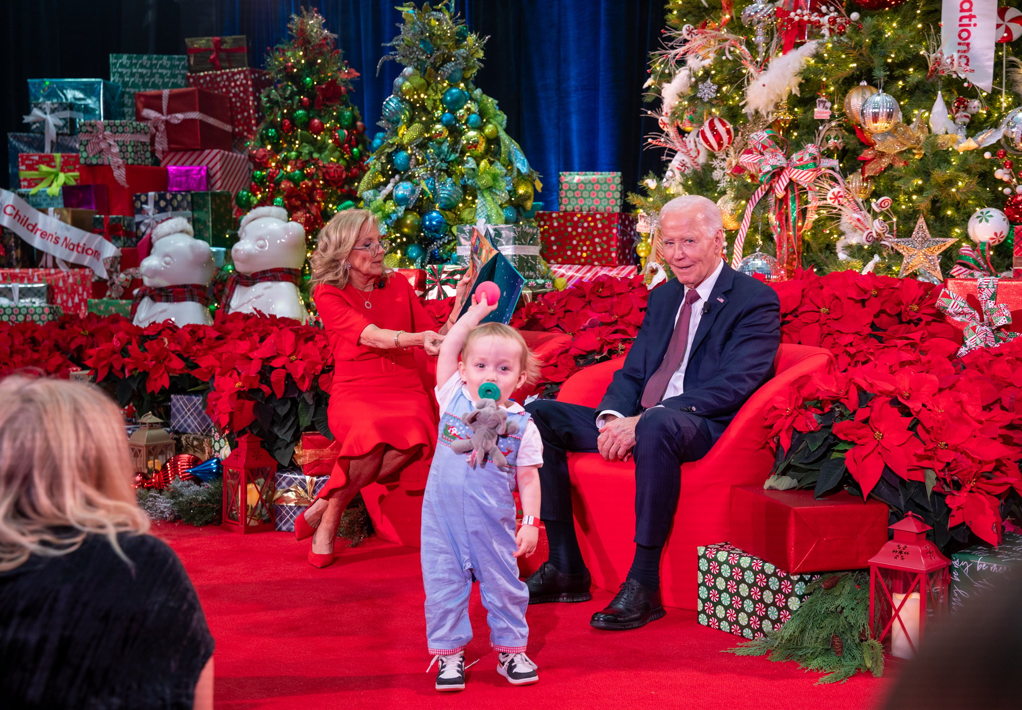 A toddler holds a ball up standing near President Joe Biden and First Lady Jill Biden while the First Lady reads 'Twas the Night Before Christmas to patients and families at Children's National Hospital