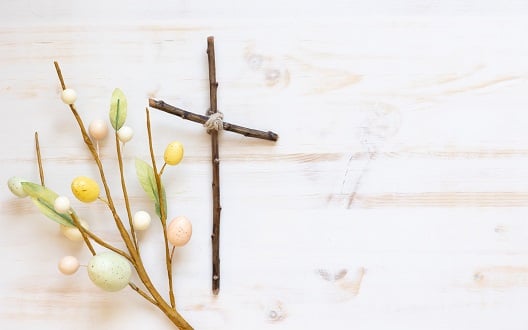 A cross made out of sticks lies next to a branch of a tree decorated with mini Easter eggs