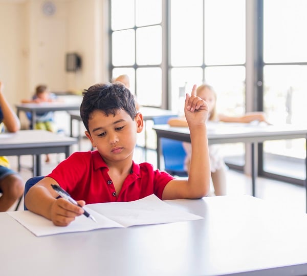 Boy in a red shirt sits at a desk with his hand raised.