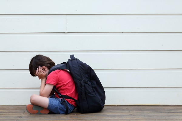 A child sits holding their head in their hands while wearing a backpack.