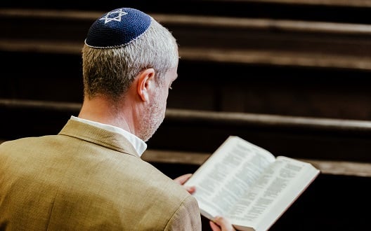 A Jewish man wearing a blue yarmulke reads a prayer book.