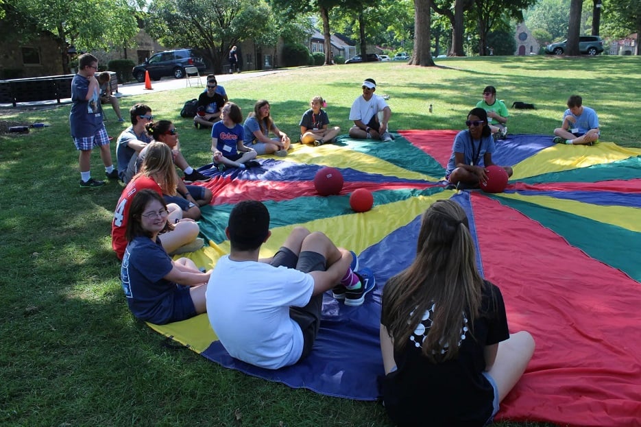 Kids sit around the edge of a colorful parachute playing a game with balls