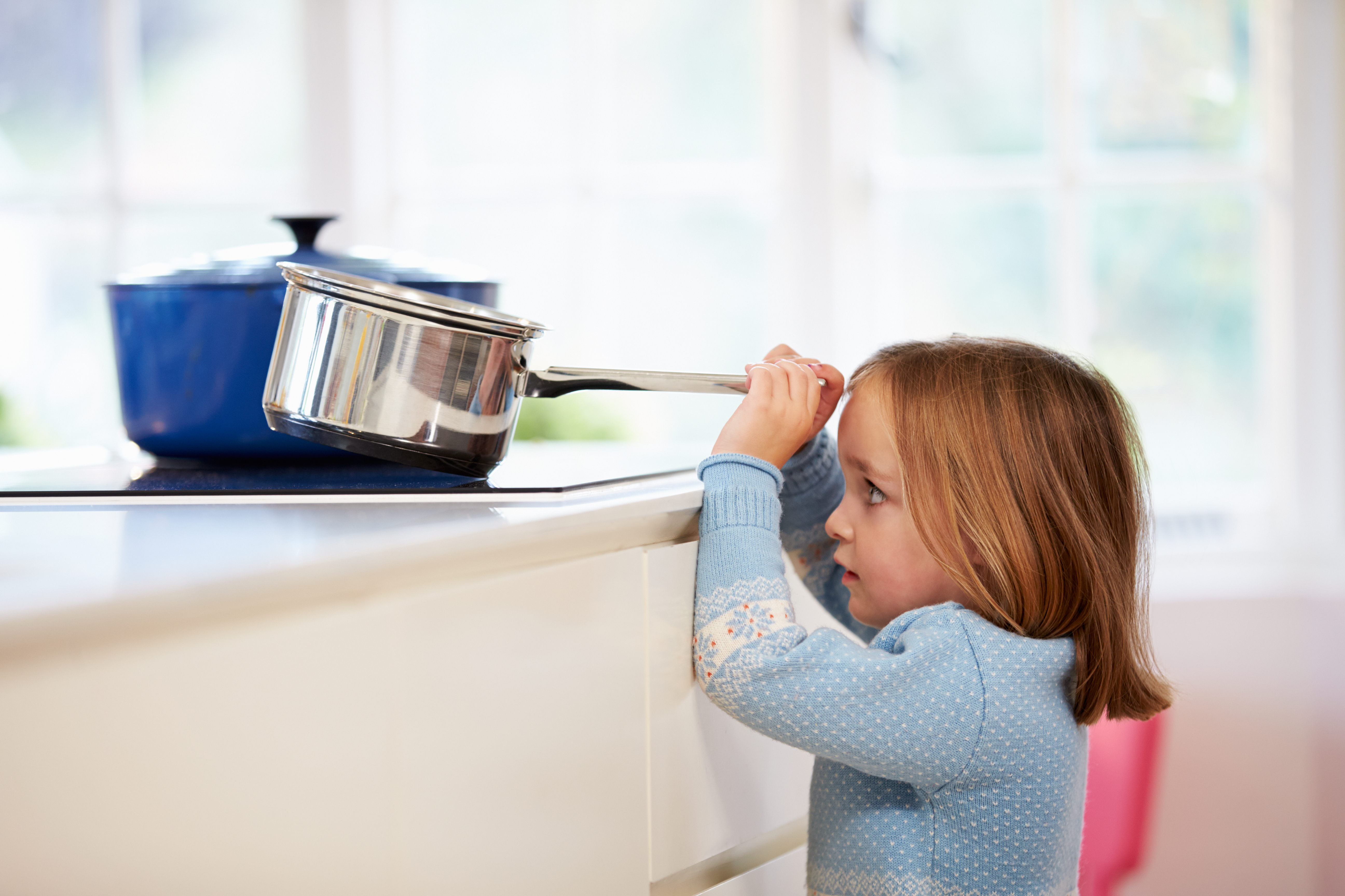 A little girl reaches to hold a pot handle on a hot stove, tilting the pot toward her.