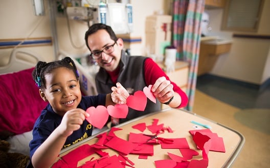 A young girl sits in her hospital bed holding red paper cut out hearts with a nurse.