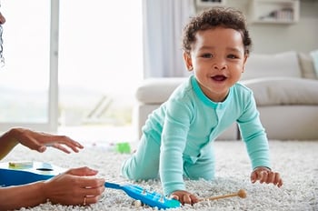 A baby boy crawls on the floor while playing with musical instruments and smiling.