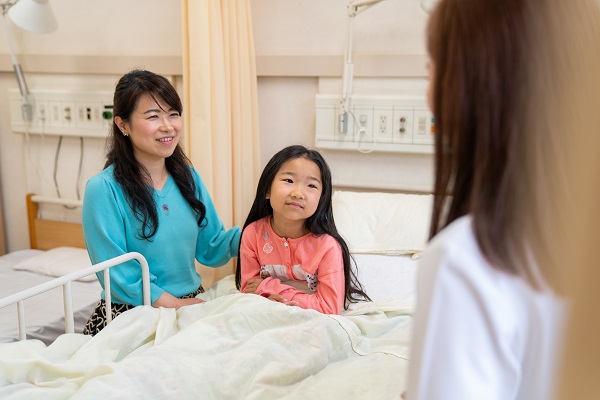 A girl sits in a hospital bed with her mother by her side as they chat with a doctor.