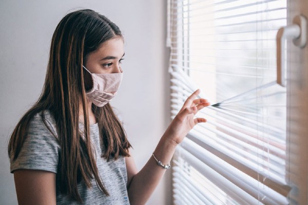 Teenage girl in mask peeking out the blinds on her window longingly