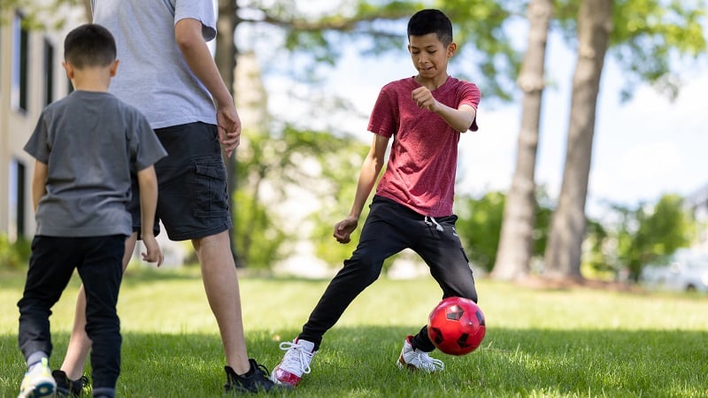 Two boys and a young man kick a red soccer ball outside