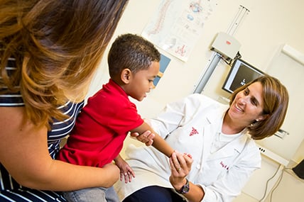 female doctor examines arm of smiling boy who sits in his mother's lap