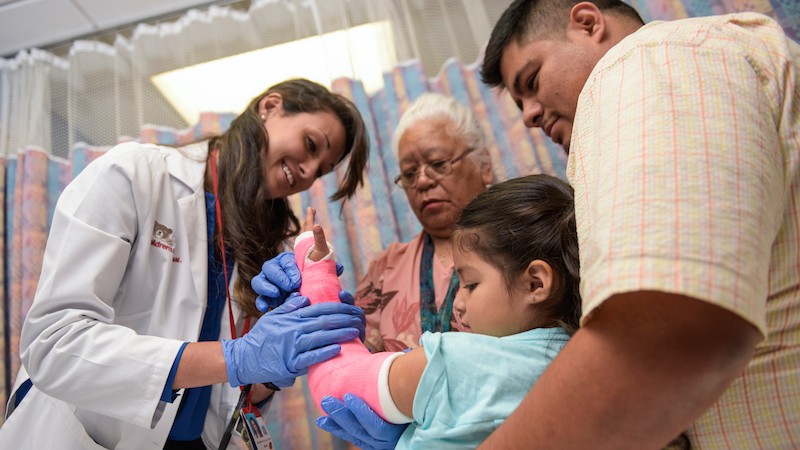 Su Visita | Doctor and nurse inspecting cast on a young girl in her father's lap