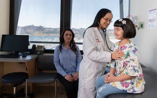 A cardiologist listens to a young girl's heart while the girl's mom looks on