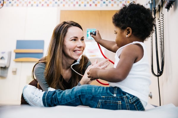 A toddler holds a stethoscope up to a smiling doctor.