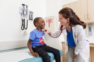 A doctor leans over to listen to a boy's heart with her stethoscope.