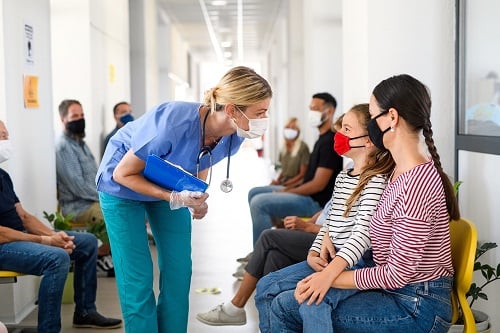 A female health care provider chats with a mother and daughter seated in a hallway.