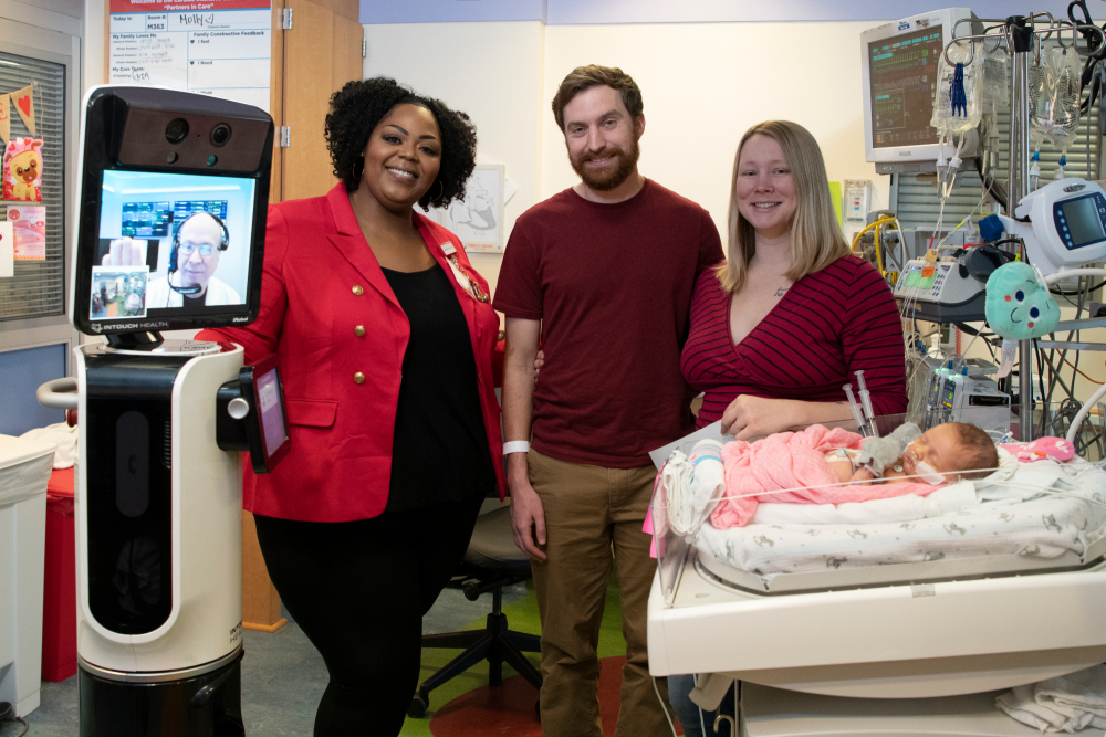 monique and dr. bear pose with parents and an infant sleeping on a hospital bed