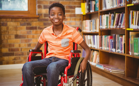 A boy sits smiling in a wheelchair in a library with bookshelves in the background.