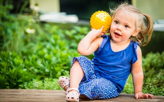 A toddler girl holds a yellow ball while sitting on a bench in the healing garden at Children's National Hospital.