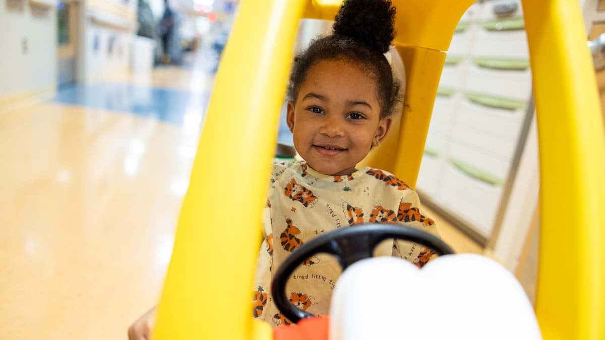 young girl driving toy car down hospital hallway