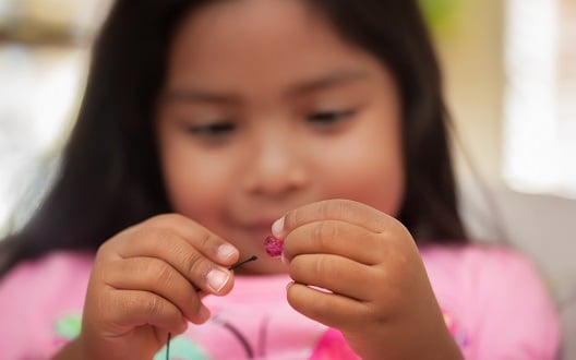 A young girl in a pink shirt carefully puts a bead on a string